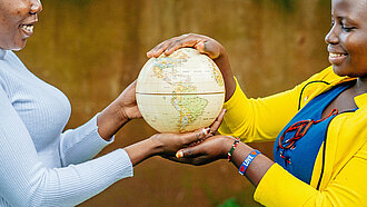 Ein Mädchen in gelber Bluse und ein Mädchen in grauem Pullover halten zusammen eine Weltkugel in ihren Händen