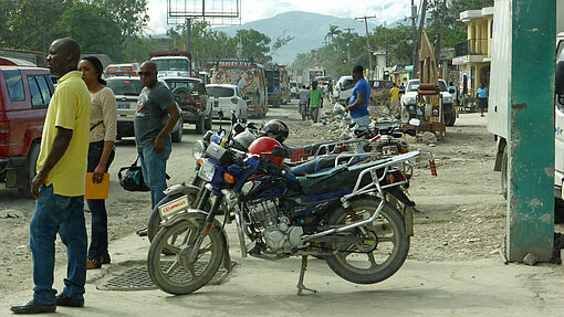 Über die verstopften Straßen von Port-au-Prince entschwindet ein Tap-Tap. Männer stehen an einer Straße neben Motorrädern.