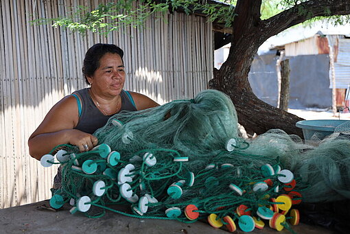 Eine Frau sitzt und repariert sorgfältig ein Fischernetz mit den Händen.