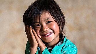 Ein junges Mädchen hält den Kopf schief und auf ihre beiden Hände gelehnt. Sie grinst in die Kamera. 