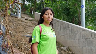 Eine junge Frau mit grünem T-Shirt steht an einem Trampelpfad im Unterholz