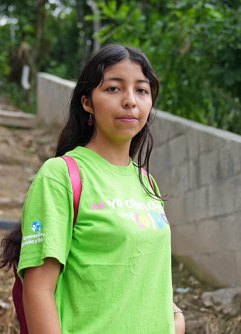 Eine junge Frau mit grünem T-Shirt steht an einem Trampelpfad im Unterholz