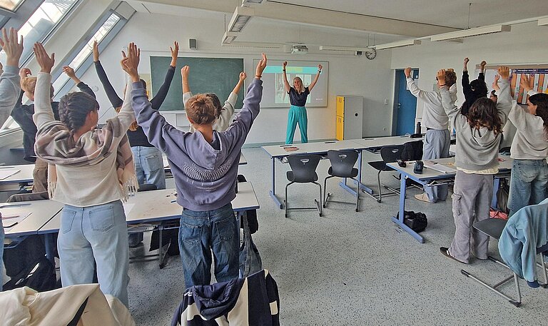 Eine Frau steht in einem Klassenzimmer vor Schulkindern und macht eine Sportübung an der Tafel vor