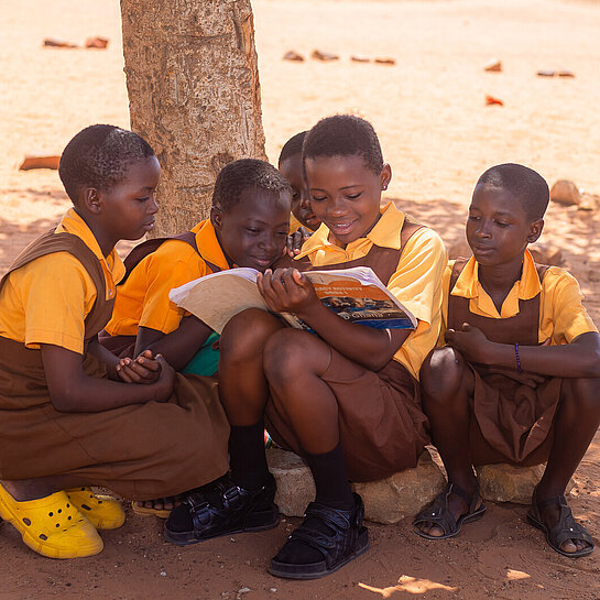 Fünf ghanaische Mädchen im Jugendalter mit Schuluniform sitzen im Schatten eines Baumes. Eines hält ein Buch in der Hand, in das alle gemeinsam reinschauen