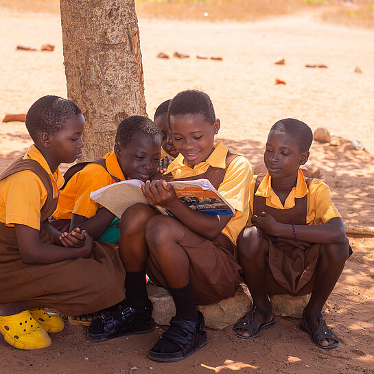 Fünf ghanaische Mädchen im Jugendalter mit Schuluniform sitzen im Schatten eines Baumes. Eines hält ein Buch in der Hand, in das alle gemeinsam reinschauen