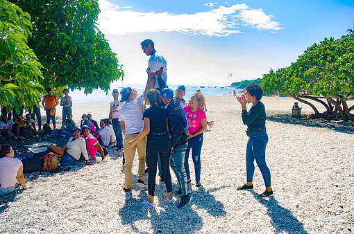Die Provinz Barahona hat wunderschöne Strände, die Tourist:innen anlocken – leider auch solche, die Jugendliche sexuell ausbeuten. Eine große Gruppe Jugendlicher ist am Strand. Einige sitzen im Schatten eines Baumes, andere stehen zusammen.