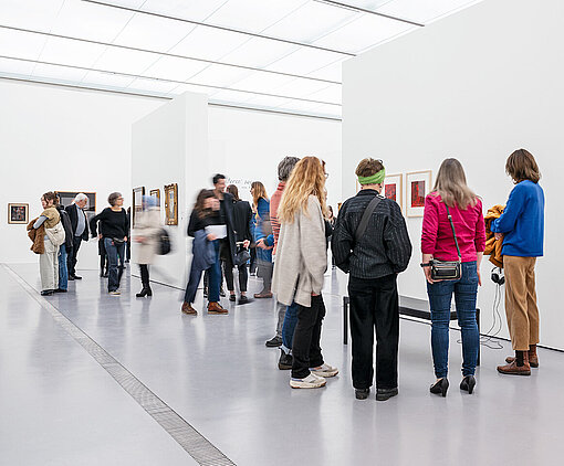 Personen stehen vor einer schneeweißen Wand, an der Fotos und bunte Ölbilder montiert sind