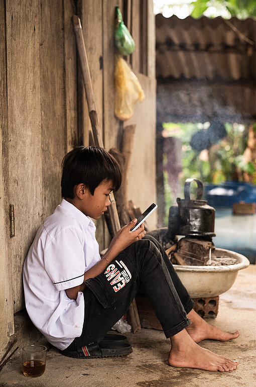 Ein Junge hockt auf einer Bastmatte und liest auf seinem Smartphone.