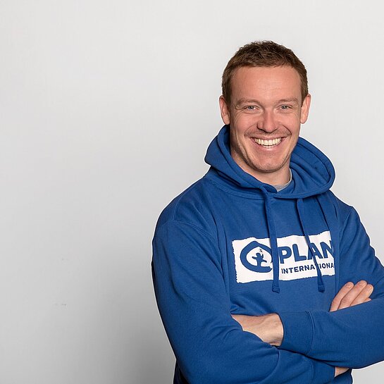 Felix Loch trägt einen Plan International-Hoddie und steht mit verschränkten Armen da. Er lächelt in die Kamera.