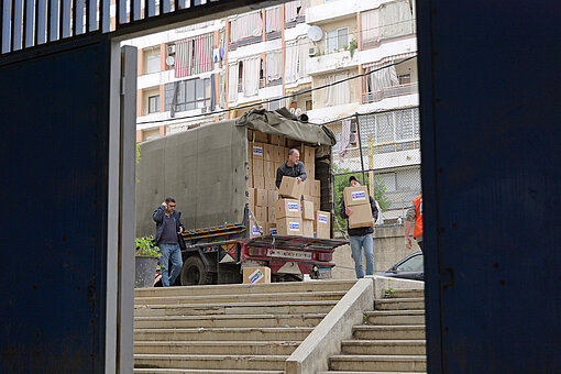 Ein Lastwagen, beladen mit Hilfspaketen von Plan International, steht vor Gebäuden in städtischer Umgebung. Mitarbeitende entladen die Kartons.
