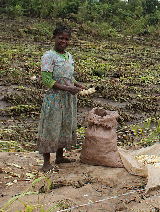Eine Frau steht vor einem überschwemmten Feld und hält einen einzelnen Maiskolben in der Hand