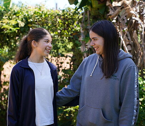 Zwei Teenager stehen in einem Garten. Eine hat ihren Arm um die andere gelegt.