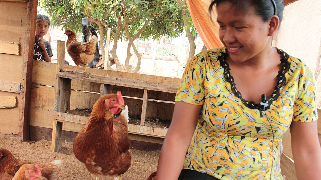 Eine Jugendliche sitzt in einem Hühnerstall und lächelt auf ein Huhn herab, das neben ihr sitzt. 