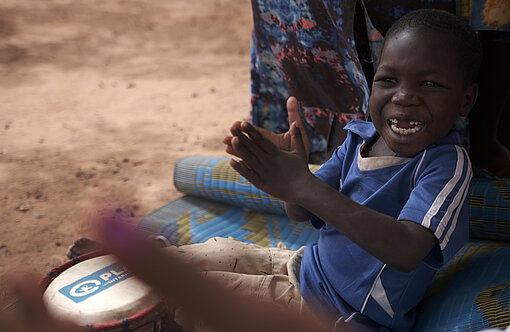 Ein Junge sitzt auf dem Boden, schlägt auf Bongo-Trommeln und schaut lachend in die Kamera