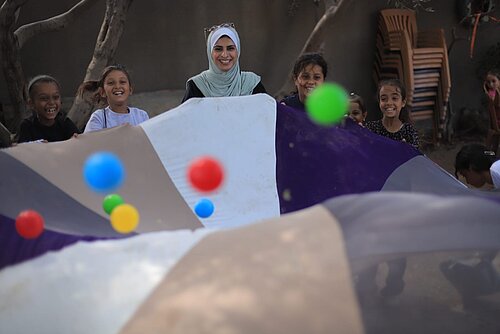 Eine Frau mit Kopftuch steht mit mehreren lachenden Kindern im Freien. Gemeinsam halten sie ein großes buntes Tuch, über dem farbige Bälle in der Luft sind.