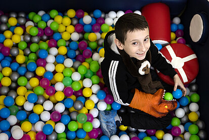 Damir, ein Junge, sitzt fröhlich im bunten Bällebad eines kinderfreundlichen Raums. Er hält ein grünes Ball und trägt eine Dino-Handschuh. Die vielen farbigen Bälle um ihn herum verleihen dem Bild eine fröhliche Atmosphäre.