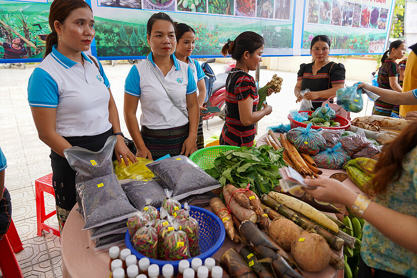 Mehrere Frauen stehen hinter einem Tisch auf einem Markt in Vietnam, auf dem verschiedene lokale Lebensmittel wie Gemüse, Gewürze und Kräuter zum Verkauf angeboten werden. Die Frauen tragen weiße Shirts mit blauen Ärmeln. Im Vordergrund ist eine Hand beim Bezahlen mit Geldscheinen zu sehen.
