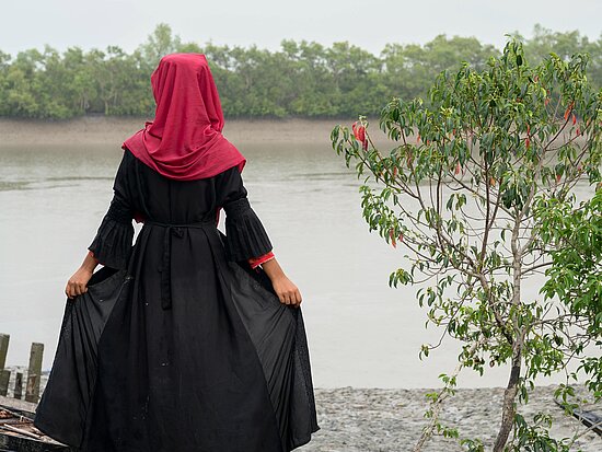 Tasnim aus Bangladesch steht in einem schwarzen Kleid mit rotem Kopftuch und schaut auf einen Fluss. Sie zeigt nur ihre Rückseite und hält mit ihren Händen ihren voluminösen Rock fest.
