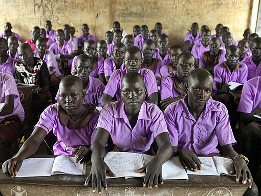 Die Hungerkrise macht den Schüler:innen im Südsudan das Lernen schwer. Viele Schüler:innen sitzen in einem Klassenraum und schauen in die Kamera. Sie tragen alle eine lila Schuluniform.