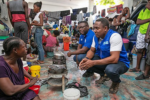 Prospery Raymond, Länderdirektor von Plan International Haiti (re.), und Programmleiter Emmanuel Michel David (2 v. r.) erkundigen sich bei geflüchteten Familien nach den Lebensumständen in der Notunterkunft Zwei Männer hocken sich zu geflüchteten Menschen