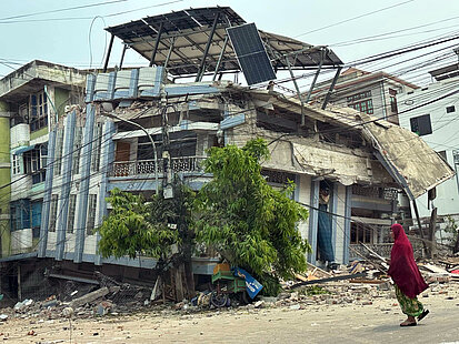 Eine Frau geht durch eine Straße in Myanmar. Hinter ihr ist ein vom Erdbeben teilweise eingestürztes Haus.