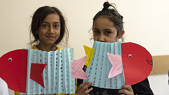 Zwei Mädchen halten Fischer aus Papier hoch, die sie gebastelt haben