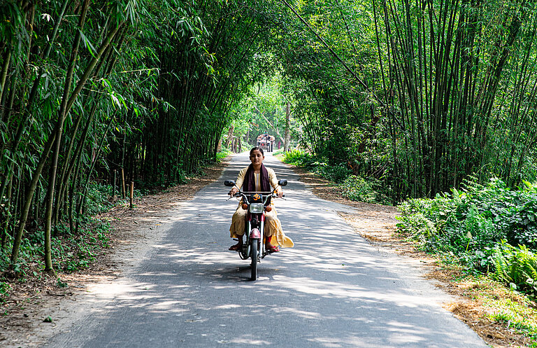Eine junge Frau aus Bangladesch fährt auf einem Moped über eine Schotterstraße