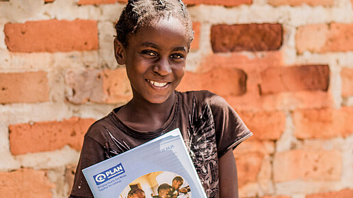 Ein Mädchen steht vor einer Mauer und hält ein Schulheft von Plan in der Hand.