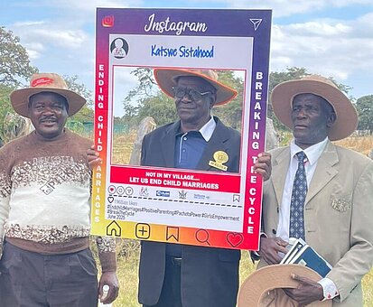 Drei Männer stehen nebeneinander und halten einen überdimensionalen Instagram-Rahmen, auf dem der Text "Not in my village, LET US END CHILD MARRIAGES" steht. Sie tragen Hüte und verschiedene Kleidungsstücke: Ein Mann trägt einen braunen Pullover, ein anderer einen schwarzen Anzug und der dritte einen beigen Anzug. Im Hintergrund ist ein ländliches Gebiet zu sehen.