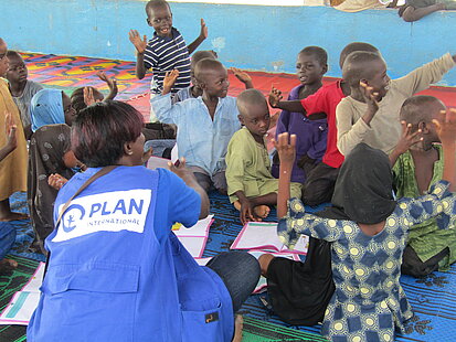 Kinder sitzen mit einer Mitarbeiterin von Plan International vor aufgeschlagenen Schulheften