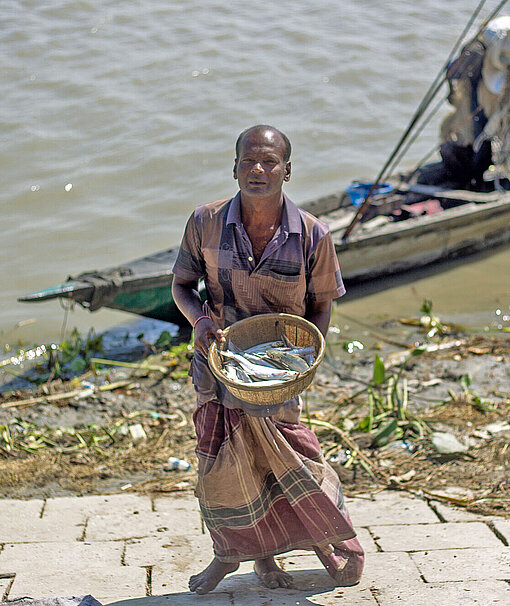 Ein Mann in einem Sarong-Wickelrock steht vor einem Fischerboot am Ufer eines Flusses