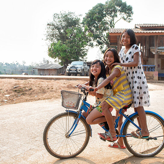 Drei Mädchen spielen sitzen auf einem Fahrrad und lachen in die Kamera.