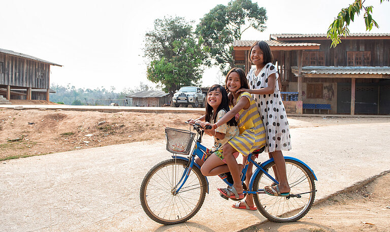 Drei Mädchen spielen sitzen auf einem Fahrrad und lachen in die Kamera.