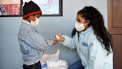 Ein kleiner Junge mit Maske bekommt von einer Plan-Mitarbeiterin Desinfektionsspray in die Hände gesprüht