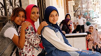 Drei Mädchen lächeln in die Kamera, im Hintergrund weitere Jugendliche auf einer Straße in Ägypten