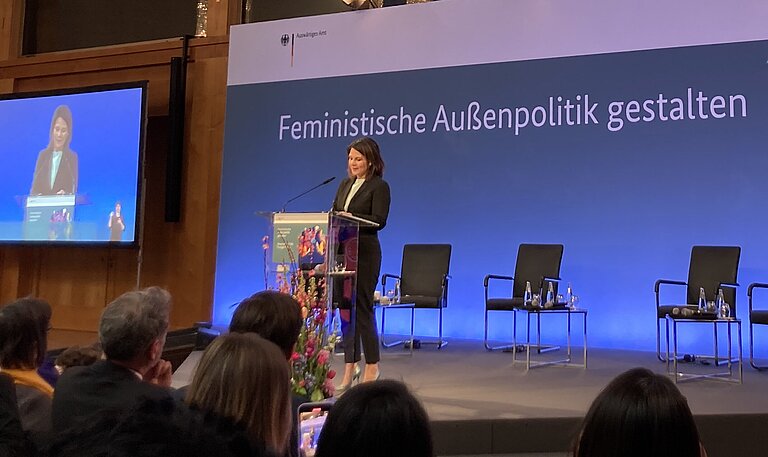 Ministerin Baerbock steht auf einer Bühne und spricht. Im Hintergrund steht der Schriftzug "Feministische Außenpolitik"