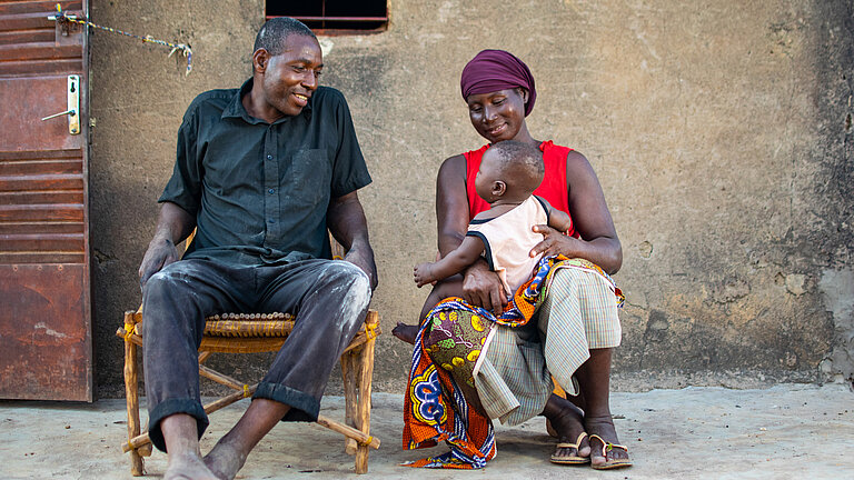 Auf einer Bank sitzen Awa (rechts) mit ihrem Baby auf dem Schoß, und Awas Mann (links) und lachen gemeinsam.