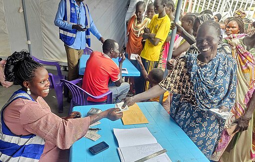 Nach der Registrierung durch das WFP erhalten bedürftige Personen aus dem Bezirk Pigi im Norden von Südsudan Bezugsscheine für Nahrungsmittelhilfe An einem Klapptisch sitzend trägt eine Frau Namen von Personen in eine Liste ein und verteilt Bezugsscheine an wartende Menschen