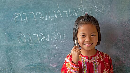 Kind steht lächelnd mit einem Kreidestück in der Hand vor einer beschriebenen Tafel