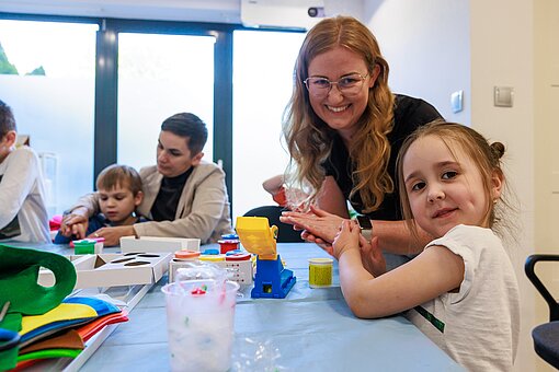 Mehrere Erwachsene sitzen mit Kindern am Tisch, auf dem Tisch liegt Material zum Basteln. Die Kinder basteln, die Erwachsenen unterstützen sie dabei.