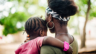 Eine junge Mutter mit einem jungen Mädchen auf dem Arm, die beiden sind von hinten zu sehen