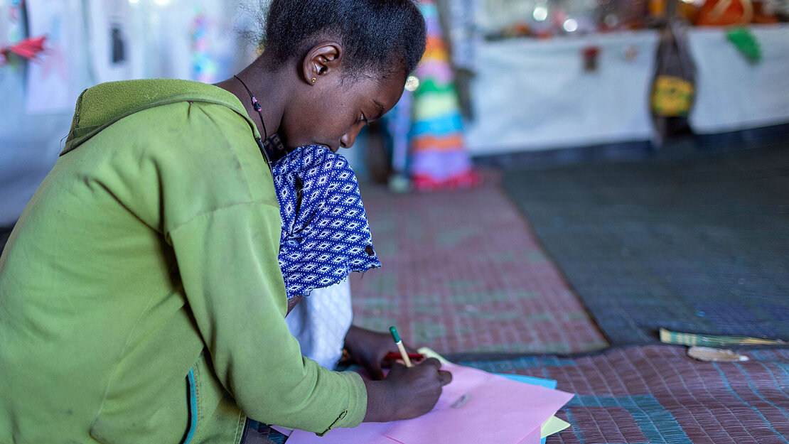 Ein Mädchen kniet auf dem Boden und malt auf ein Blatt Papier.