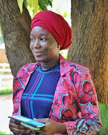Mama Sampy berichtet, wie sich Nahrungsmittelknappheit und Armut auf das Leben von Mädchen in Mali auswirken. Mama Sampy steht vor einem Baum. Sie trägt einen bunten Blazer und ein rotes Kopftuch.