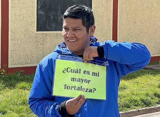 Ein Mann in blauer Jacke hält ein Schild mit der Aufschrift "cuál es mi mayor fortaleza?" hoch.