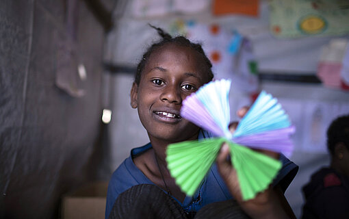 Ein Mädchen hält eine gebastelte Blume in die Kamera. Im Hintergrund hängen bunte Bilder an der Wand.