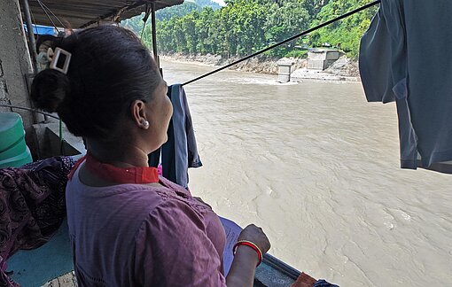 Eine Frau in pinker Bekleidung steht auf einem Balkon und blickt auf einen Fluss mit grauem Wasser, in dem die Reste einer Brücke stehen.