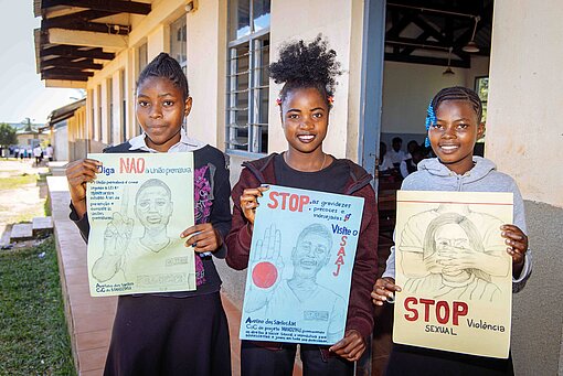 Mädchen in Mosambik organisieren sich im Plan-Jugendclub „Champions of Change“ und machen unter anderem mit selbst gestalteten Plakaten auf die Folgen von Missbrauch, Gewalt und sexuellen Übergriffen aufmerksam Drei Mädchen halten selbst gestaltete Wandplakate in die Kamera