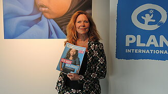 Marion Kracht steht in einem Raum. Im Hintergrund hängt ein großes Bild von einem kleinen Mädchen und das Logo von Plan International