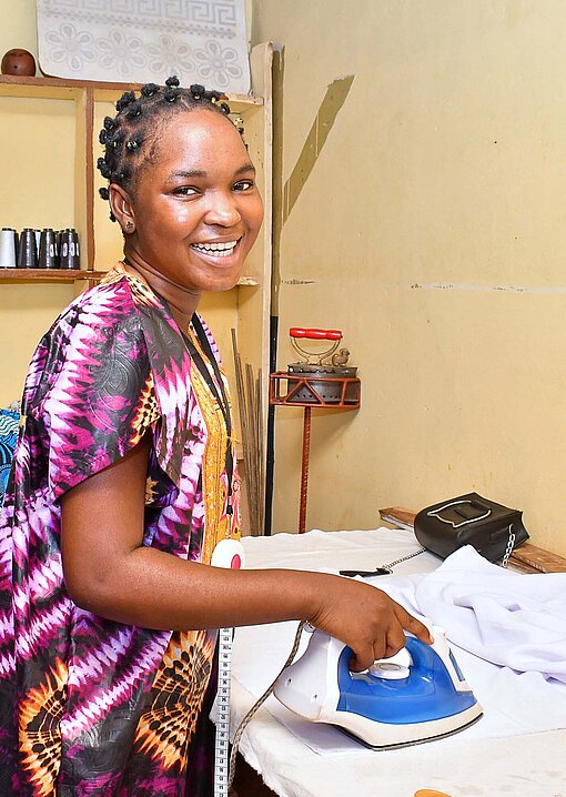 Eine Frau in buntem Kleid steht an einem Tisch und bügelt einen Stoff