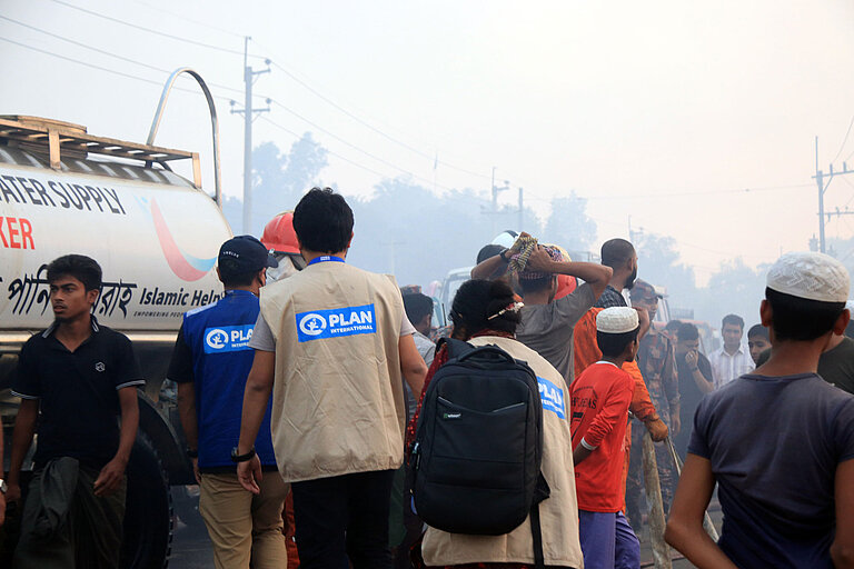 Drei Menschen mit Plan-Westen laufen durch eine Menschenmenge. Im Hintergrund steht ein Wasser-Transporter. In der Luft liegt Rauch.